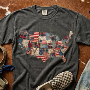 May include: Charcoal grey Comfort Colors t-shirt with a patchwork map of the USA. The map is made from fabric patches with American flag and patriotic designs. A pair of checkered Vans shoes is visible.