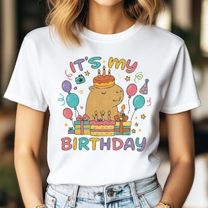 Pode incluir: Camiseta branca com um gráfico colorido que diz "It's my Birthday". O gráfico apresenta uma capivara de desenho animado com um bolo de aniversário, balões, presentes e confetes. A camiseta é feita de um material macio.