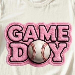 May include: Cream-colored t-shirt featuring a pink, fluffy "GAME DAY" design with black outlines. A baseball is centered within the design. The baseball has red stitching and a worn appearance.