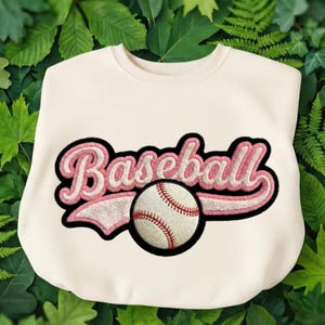 Puede incluir: Sudadera de color crema con la palabra "Baseball" en rosa, contorneada en negro, y un gráfico de béisbol. La pelota de béisbol es blanca con costuras rojas y un contorno negro. Prenda de vestir deportiva informal.