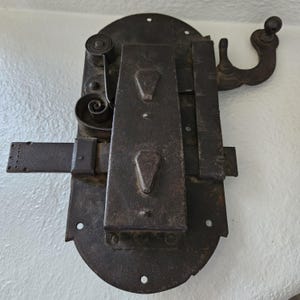 May include: A vintage, dark brown metal lock with intricate details. The lock features a spring mechanism, a sliding bolt, and a decorative handle. The metal has a weathered appearance, suggesting age and use. The lock is mounted on a white surface.