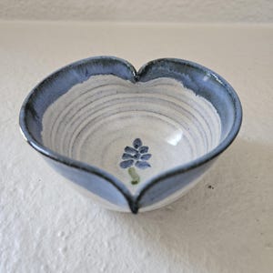 May include: A heart-shaped ceramic bowl with a blue rim and a floral design. The bowl's interior is white with blue and gray concentric lines. A blue floral design with green stems is centered in the bowl.
