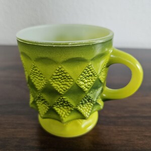 May include: A vintage green glass mug with a textured diamond pattern. The mug has a rounded base and a handle. The interior is a lighter green color. The mug is likely from the mid-20th century.