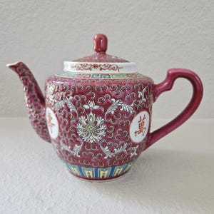 May include: A maroon and white porcelain teapot with a floral design and Chinese characters. The teapot has a curved spout, a loop handle, and a lid with a small knob. The design includes a white floral center and a decorative border.