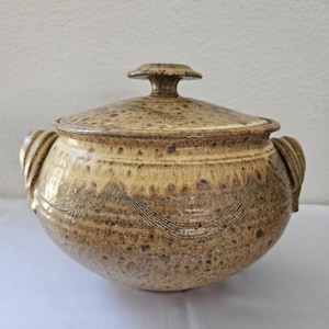 May include: A handcrafted ceramic casserole dish with a lid. The dish is a speckled tan color with brown accents and a decorative pattern around the middle. It has two handles and a knob on the lid.