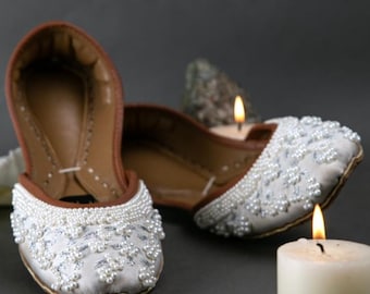 May include: A pair of white and silver embellished slip-on shoes with a brown leather interior and trim. The shoes are decorated with small pearls and floral designs. The image is set against a dark background with lit candles.