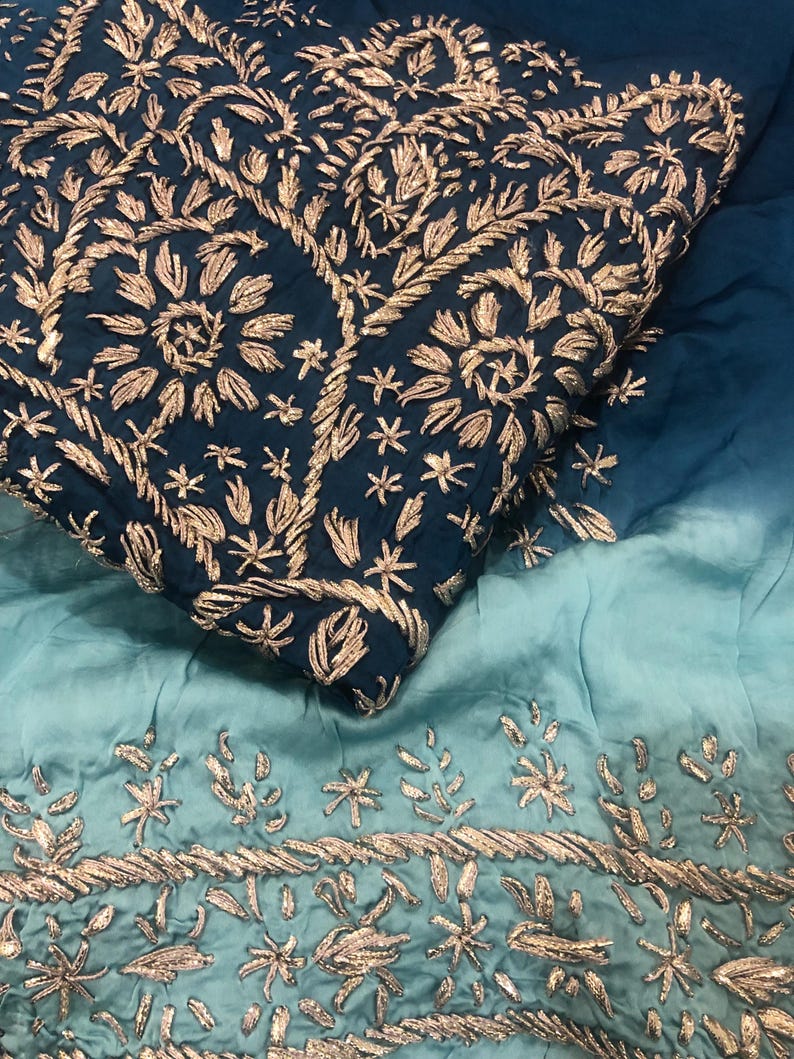 May include: A fabric textile featuring an ombre gradient from deep navy blue to light teal. The navy section is densely embroidered with metallic gold floral and vine patterns, while the lighter teal section features a horizontal border of gold.