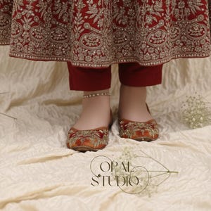 May include: A pair of orange velvet flats featuring intricate gold embroidery and beadwork, worn with deep red trousers and a long, matching red skirt with heavy gold floral embroidery. The text OPAL STUDIO is visible in the lower center.