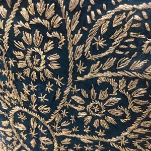 May include: A deep teal fabric featuring intricate gold metallic thread embroidery. The design consists of repeating floral and leaf motifs arranged in a dense, swirling pattern that covers the entire surface of the dark blue-green textile.