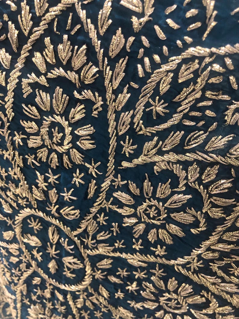 May include: A deep teal fabric featuring intricate gold metallic thread embroidery. The design consists of repeating floral and leaf motifs arranged in a dense, swirling pattern that covers the entire surface of the dark blue-green textile.