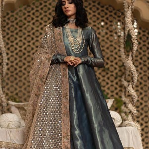 May include: A long, flowing teal dress with long sleeves. The dress is paired with a sheer, tan shawl embellished with sequins and embroidery. The model is wearing layered pearl necklaces and a large ring.