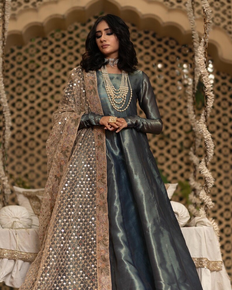 May include: A long, flowing teal dress with long sleeves. The dress is paired with a sheer, tan shawl embellished with sequins and embroidery. The model is wearing layered pearl necklaces and a large ring.