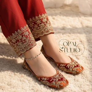 May include: Deep red trousers featuring intricate gold embroidery along the hem, paired with matching deep red flat shoes adorned with detailed gold floral embroidery. A delicate silver-toned beaded anklet is worn on the left ankle.