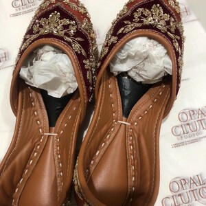 May include: A pair of slip-on flats featuring deep maroon velvet uppers adorned with intricate gold metallic embroidery and beadwork. The interior is lined with tan leather, and the shoes contain white tissue paper inserts for shape retention.