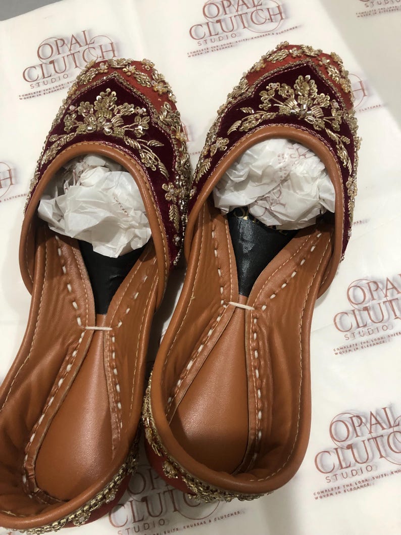 May include: A pair of slip-on flats featuring deep maroon velvet uppers adorned with intricate gold metallic embroidery and beadwork. The interior is lined with tan leather, and the shoes contain white tissue paper inserts for shape retention.