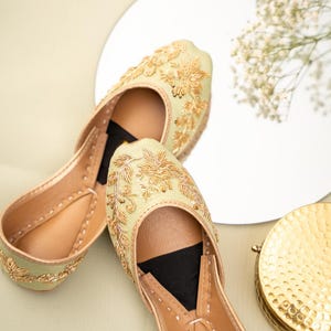 May include: A pair of light green and gold embroidered flats. The shoes feature intricate gold floral embroidery on the upper and a tan leather sole. The flats are displayed on a light green surface with a mirror and gold decorative item.