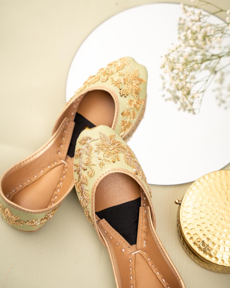 May include: A pair of light green and gold embroidered flats. The shoes feature intricate gold floral embroidery on the upper and a tan leather sole. The flats are displayed on a light green surface with a mirror and gold decorative item.