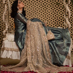 May include: A woman in a teal-colored dress with a sheer overlay, seated on a swing. A gold-colored, sequined shawl drapes over the swing. The background features a patterned wall and the floor is covered in red flower petals.