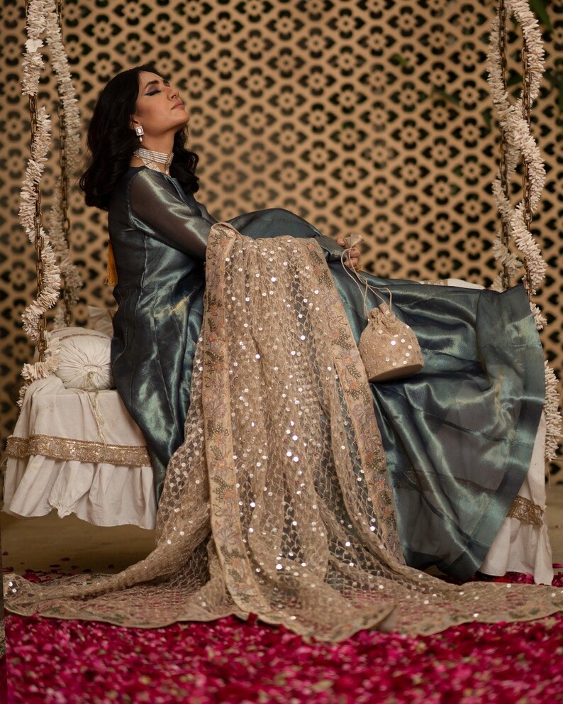 May include: A woman in a teal-colored dress with a sheer overlay, seated on a swing. A gold-colored, sequined shawl drapes over the swing. The background features a patterned wall and the floor is covered in red flower petals.