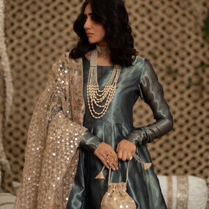 May include: A woman wearing a teal-colored, long-sleeved dress with a layered pearl necklace and a gold sequined shawl. She holds a small, gold sequined drawstring bag and wears a large, ornate ring. The dress has a flowing skirt.
