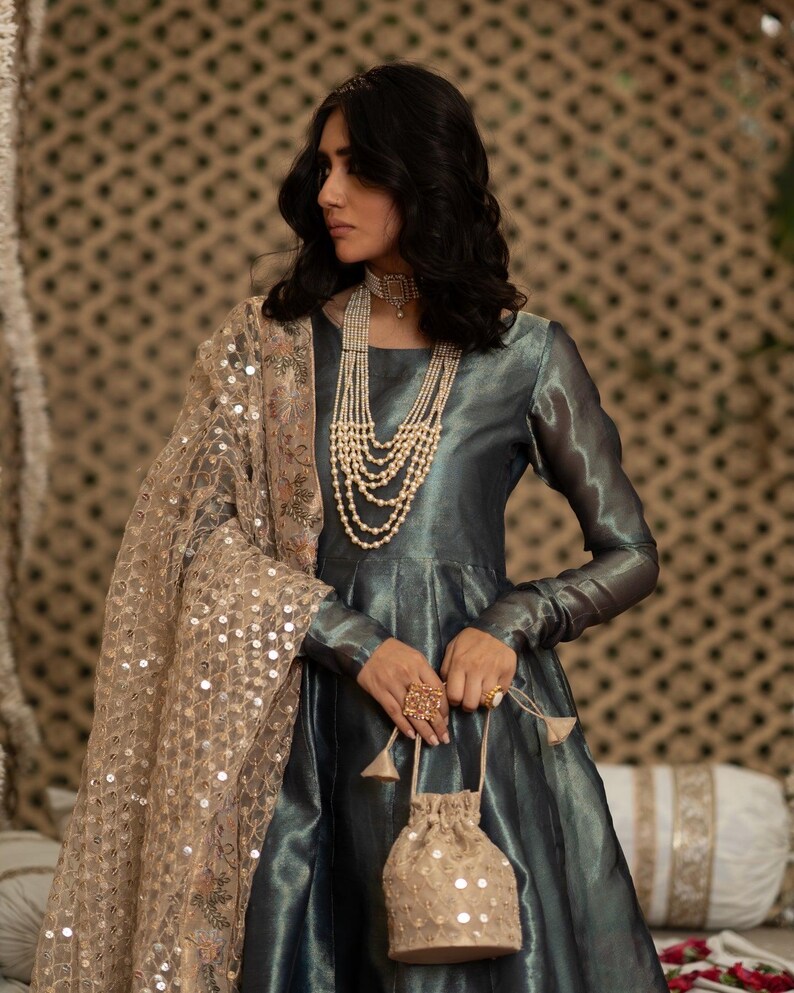 May include: A woman wearing a teal-colored, long-sleeved dress with a layered pearl necklace and a gold sequined shawl. She holds a small, gold sequined drawstring bag and wears a large, ornate ring. The dress has a flowing skirt.