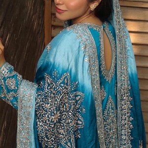 May include: A vibrant turquoise and sky blue ombre tunic featuring intricate silver metallic embroidery in floral and geometric patterns. The garment includes long sleeves, a teardrop-shaped back cutout, and a matching draped shawl overlay.