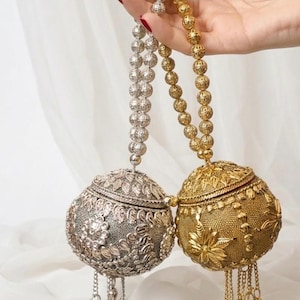 May include: Two spherical metal handbags, one silver and one gold, featuring intricate embossed floral patterns and textured surfaces. Each bag has a beaded handle and a dangling tassel of small metal bells attached to the bottom.