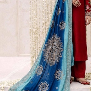 May include: A deep red long-sleeved tunic and matching trousers paired with a long, flowing dupatta. The dupatta features an ombre design transitioning from dark blue to light blue, adorned with intricate gold floral embroidery and borders.