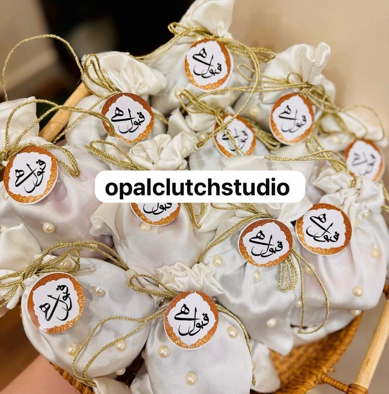 Peut inclure: Plusieurs petits sacs cadeaux en satin blanc, nou&eacute;s avec de la ficelle dor&eacute;e et orn&eacute;s d'&eacute;tiquettes rondes avec de la calligraphie arabe. Les sacs sont d&eacute;cor&eacute;s de perles et dispos&eacute;s dans un panier en osier. Le texte "opalclutchstudio" est visible.