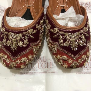 May include: A pair of maroon velvet flats featuring intricate gold embroidery with metallic thread, sequins, and small white beads. The shoes have a pointed toe, a tan leather interior lining, and a slip-on design with a structured, rounded opening.