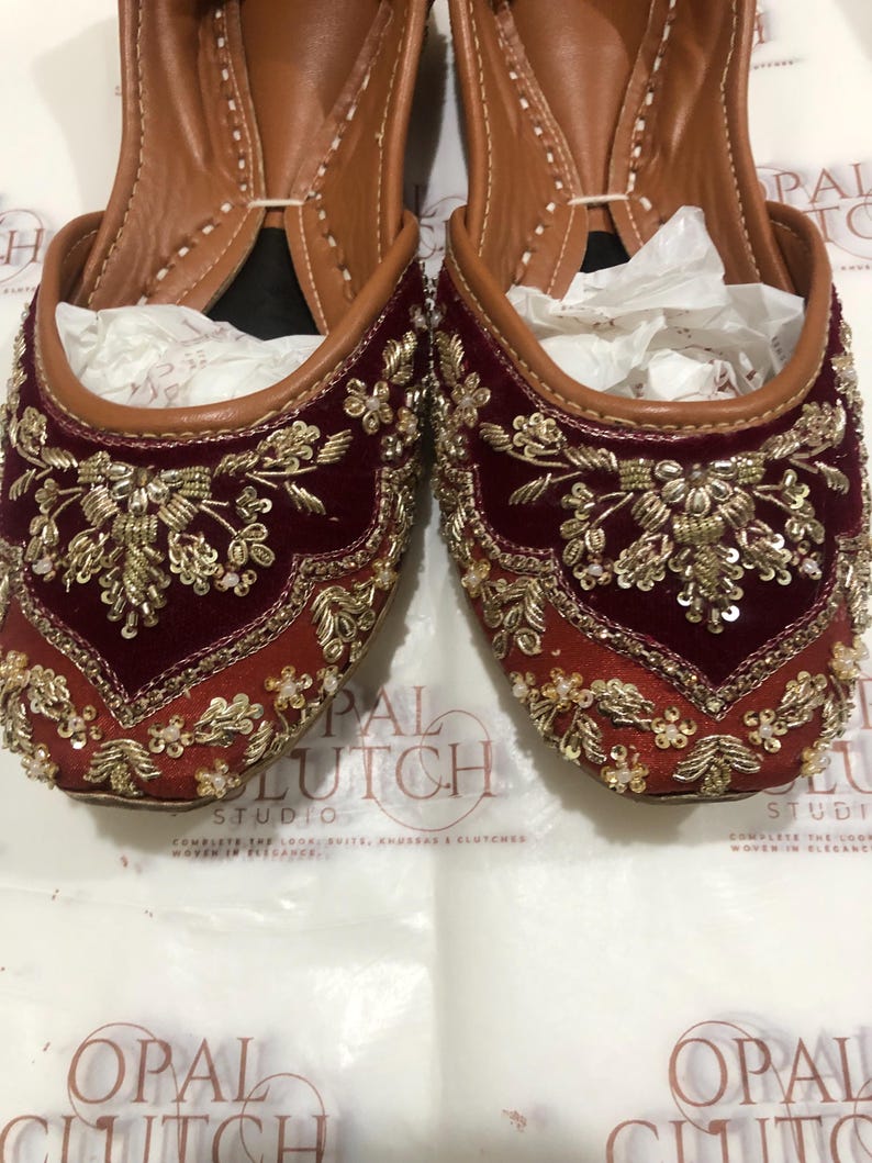May include: A pair of maroon velvet flats featuring intricate gold embroidery with metallic thread, sequins, and small white beads. The shoes have a pointed toe, a tan leather interior lining, and a slip-on design with a structured, rounded opening.