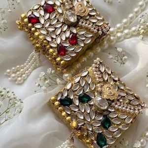 Puede incluir: Dos piezas de joyería doradas, de forma cuadrada y adornadas. Cada una está incrustada con piedras claras en forma de lágrima, gemas rojas o verdes y perlas. Cada pieza tiene una borla de perlas. Flores blancas y un collar de perlas en el fondo.