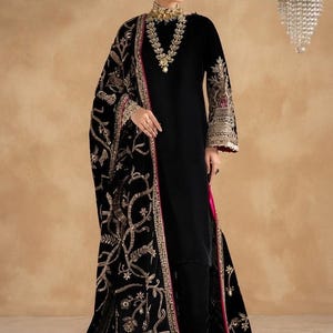 May include: A black velvet long-sleeved tunic and matching trousers paired with a black velvet shawl. The shawl and sleeves feature intricate gold embroidery and sequin detailing. The ensemble includes a vibrant pink trim along the shawl edges.