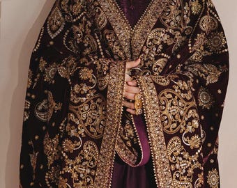 May include: A deep plum velvet shawl featuring intricate gold embroidery in paisley and floral patterns, accented with small reflective embellishments. The shawl is draped over a matching dark purple garment, showcasing a wide, heavily detailed border.
