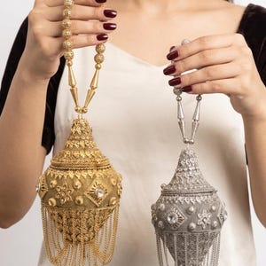May include: Two ornate, pear-shaped metal handbags, one gold and one silver, featuring intricate filigree patterns, embedded clear stones, and dangling chain fringe. Each bag has a matching beaded handle with conical accents at the base.