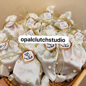 Peut inclure: Plusieurs pochettes en satin blanc, orn&eacute;es de ficelle dor&eacute;e et de petites perles. Chaque pochette est surmont&eacute;e d'une &eacute;tiquette ronde avec de la calligraphie arabe et une bordure dor&eacute;e &agrave; paillettes. Le texte "opalclutchstudio" est affich&eacute;.