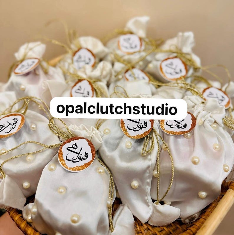 Peut inclure: Plusieurs pochettes en satin blanc, orn&eacute;es de ficelle dor&eacute;e et de petites perles. Chaque pochette est surmont&eacute;e d'une &eacute;tiquette ronde avec de la calligraphie arabe et une bordure dor&eacute;e &agrave; paillettes. Le texte "opalclutchstudio" est affich&eacute;.