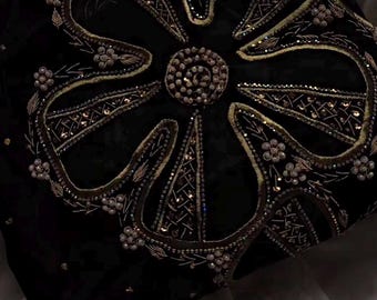 Puede incluir: Textil negro con un diseño floral bordado. El motivo central es una flor grande y estilizada con detalles intrincados en hilo dorado y plateado, rodeada de pequeños acentos florales. La tela tiene una textura aterciopelada.
