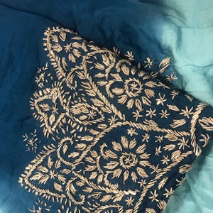 May include: A fabric textile featuring an ombre gradient transitioning from deep teal to light aqua blue. The material is adorned with intricate metallic gold thread embroidery in floral and leaf patterns, creating a textured, decorative surface.