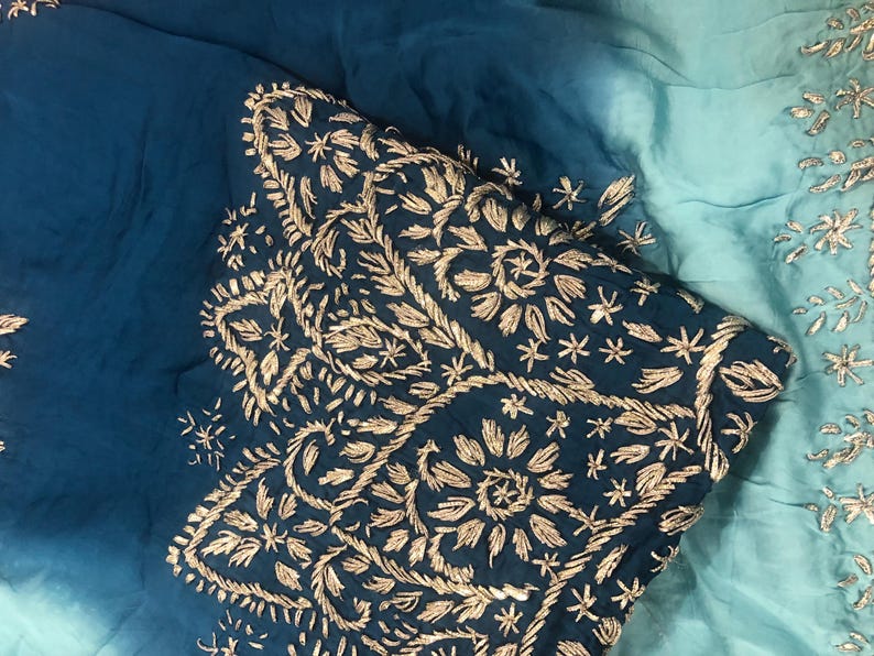 May include: A fabric textile featuring an ombre gradient transitioning from deep teal to light aqua blue. The material is adorned with intricate metallic gold thread embroidery in floral and leaf patterns, creating a textured, decorative surface.