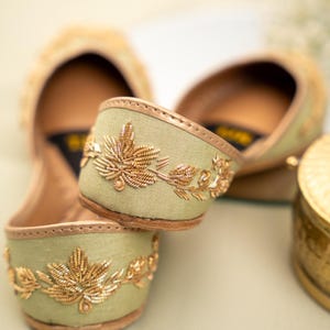 May include: Close-up of two pairs of sage green flats with gold leaf embroidery. The shoes have a rounded toe and a gold trim. The embroidery features detailed gold leaves and vines. The background is a soft, blurred neutral color.