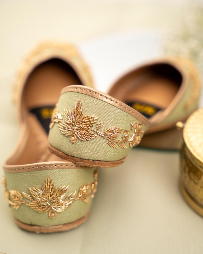 May include: Close-up of two pairs of sage green flats with gold leaf embroidery. The shoes have a rounded toe and a gold trim. The embroidery features detailed gold leaves and vines. The background is a soft, blurred neutral color.