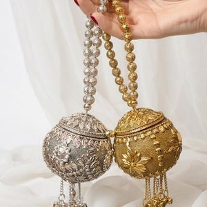 May include: Two ornate, spherical metal purses, one silver and one gold, are suspended from beaded chains. The silver purse features floral and leaf patterns, while the gold one has a textured surface with flower designs. Both have dangling charms.
