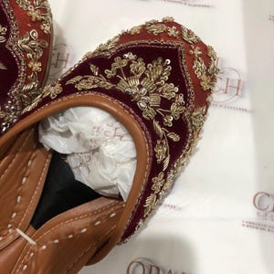 May include: A pair of deep red velvet flats featuring intricate gold embroidery, metallic thread work, and small clear beads. The shoes have a tan leather trim, a pointed toe, and a tan leather interior lining with white stitching along the edges.