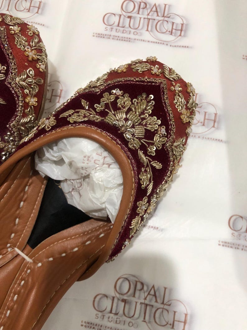 May include: A pair of deep red velvet flats featuring intricate gold embroidery, metallic thread work, and small clear beads. The shoes have a tan leather trim, a pointed toe, and a tan leather interior lining with white stitching along the edges.