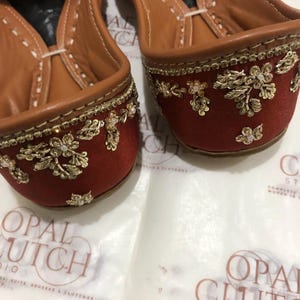 May include: A pair of rust-colored flats featuring intricate gold metallic embroidery and small white bead accents. The interior is lined with tan leather, and the shoes are positioned on a white surface with the text Opal Clutch Studio visible.