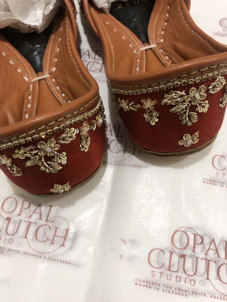 May include: A pair of rust-colored flats featuring intricate gold metallic embroidery and small white bead accents. The interior is lined with tan leather, and the shoes are positioned on a white surface with the text Opal Clutch Studio visible.
