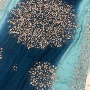 May include: A fabric textile featuring an ombre gradient from deep navy blue to light turquoise. The surface is decorated with intricate silver-toned embroidery in large circular mandala-style patterns and a detailed border along the edge.
