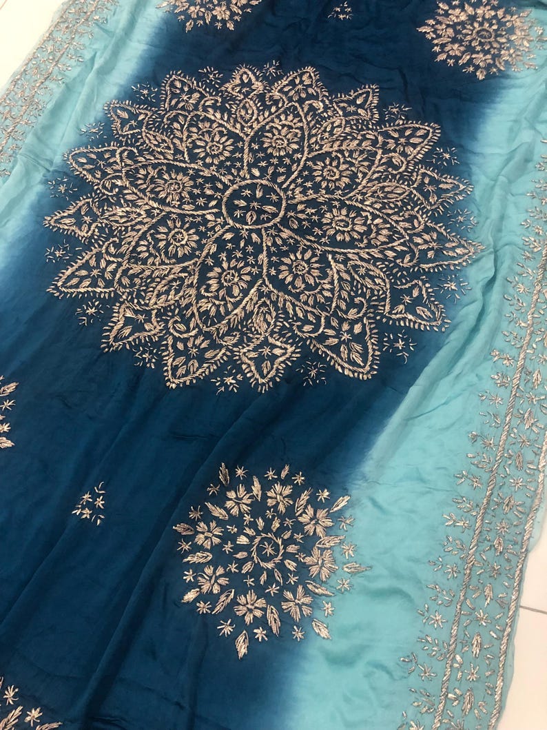 May include: A fabric textile featuring an ombre gradient from deep navy blue to light turquoise. The surface is decorated with intricate silver-toned embroidery in large circular mandala-style patterns and a detailed border along the edge.