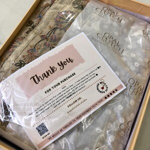 May include: A beige box contains a folded textile with floral embroidery, tissue paper with a logo, and a thank you card. The card has text that reads "Thank you" and "For your purchase". The box is on a marble surface.